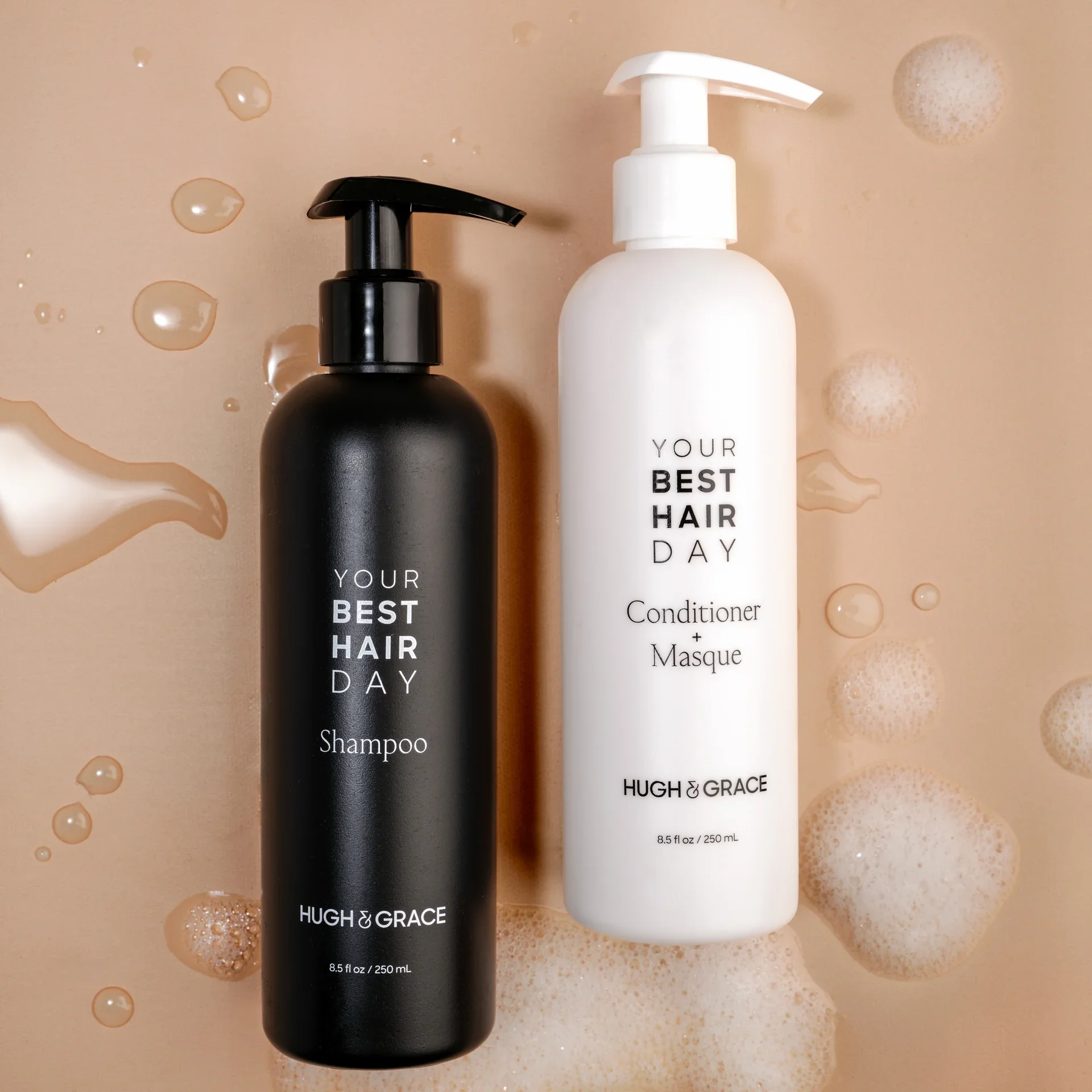 A black shampoo bottle and a white conditioner plus masque bottle on a beige surface with soap bubbles.
