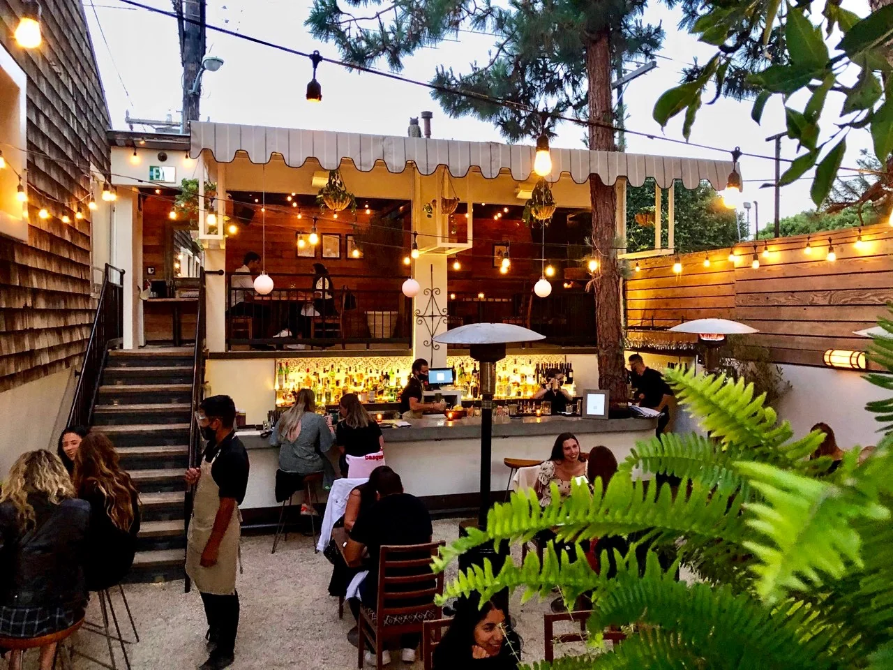 An outdoor dining area at a restaurant with hanging lights, people seated at tables, and a bar in the background.