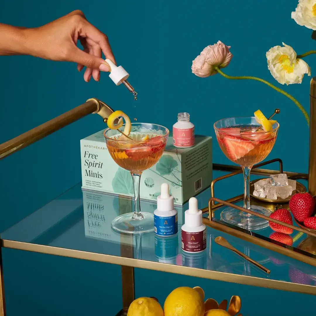 A hand drops a tincture into a citrus cocktail beside bottles of herbal wellness drops, fresh fruit, and flowers on a gold and glass bar cart.