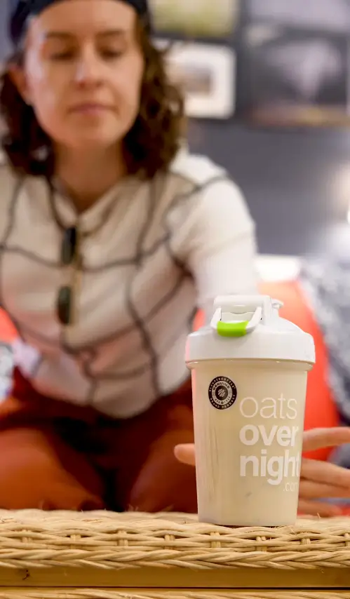 Woman reaching for an Oats Overnight shaker bottle on a wicker table in a casual indoor setting.