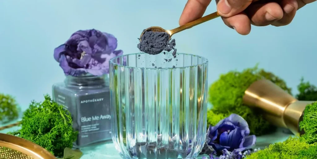 A hand holds a spoon with a blue powder over a clear glass next to a jar labeled Blue Me Away by Apothékary.
