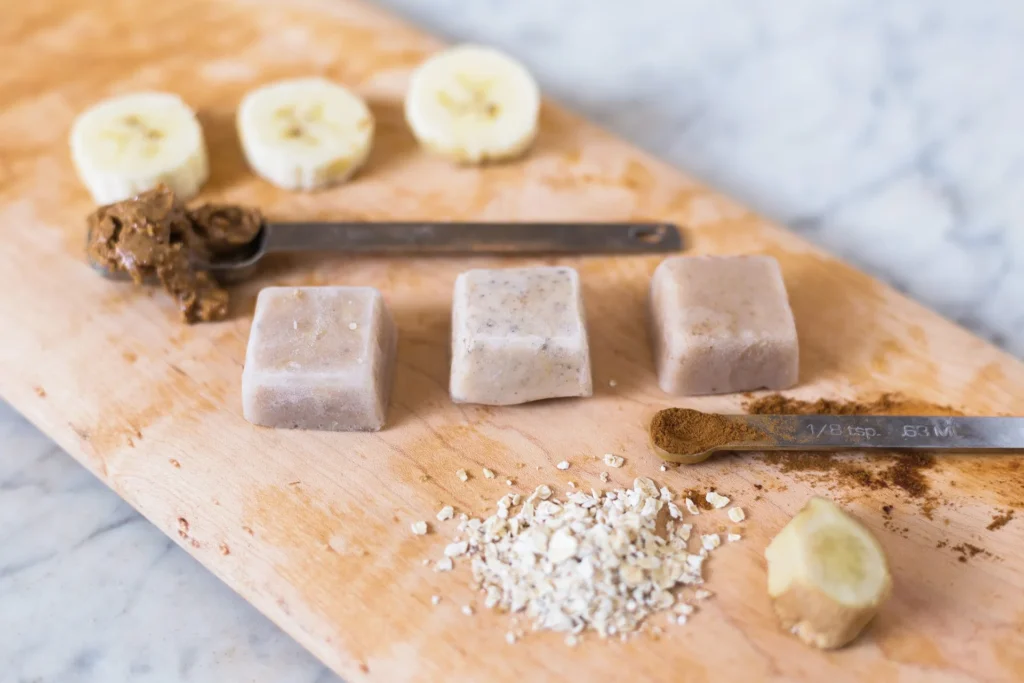 Various food ingredients arranged on a wooden surface including banana slices, nut butter, frozen cubes, oats, ginger, and cinnamon.