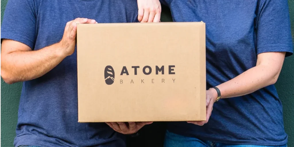 A person holding a cardboard box with the Atome Bakery logo.