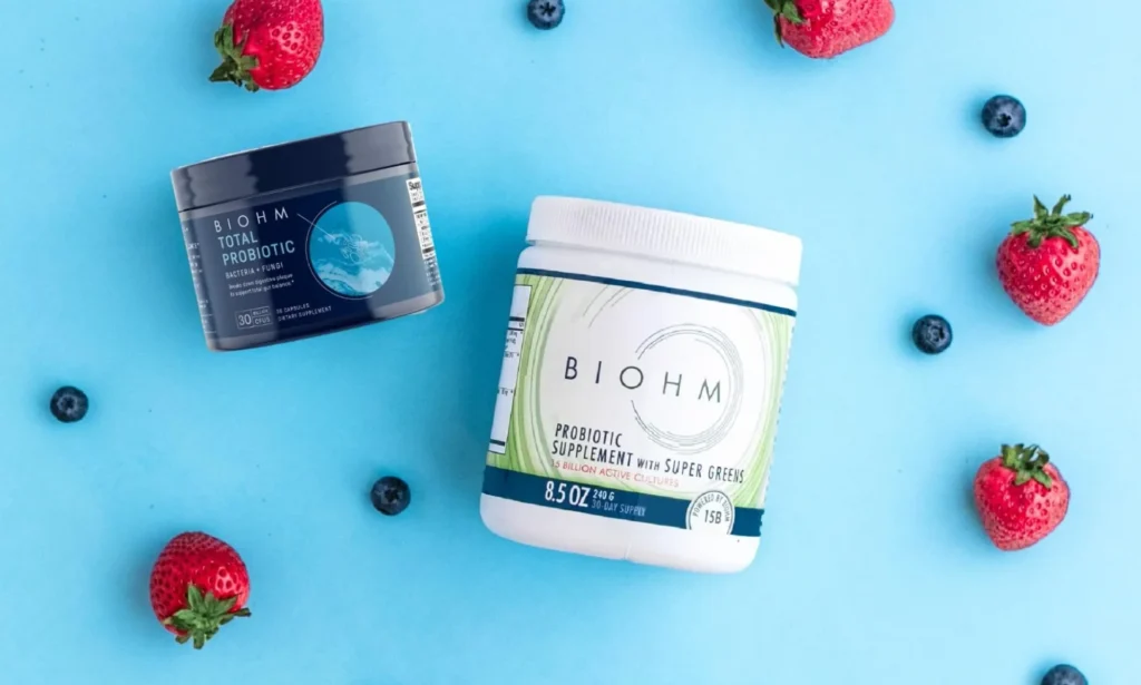 BIOHM probiotic supplements placed on a blue background surrounded by strawberries and blueberries.