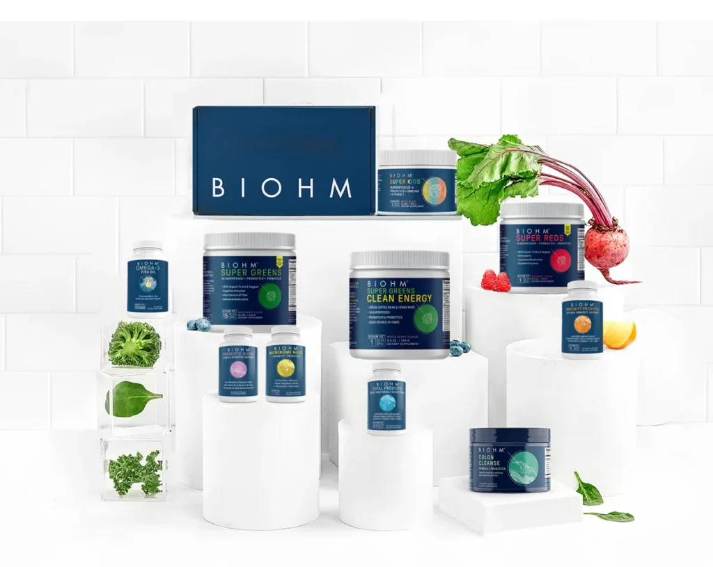 An assortment of BIOHM products displayed on stacked white pedestals with fresh vegetables.