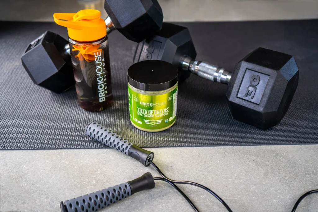 Black dumbbells, a water bottle, and a container of Field of Greens on a mat.
