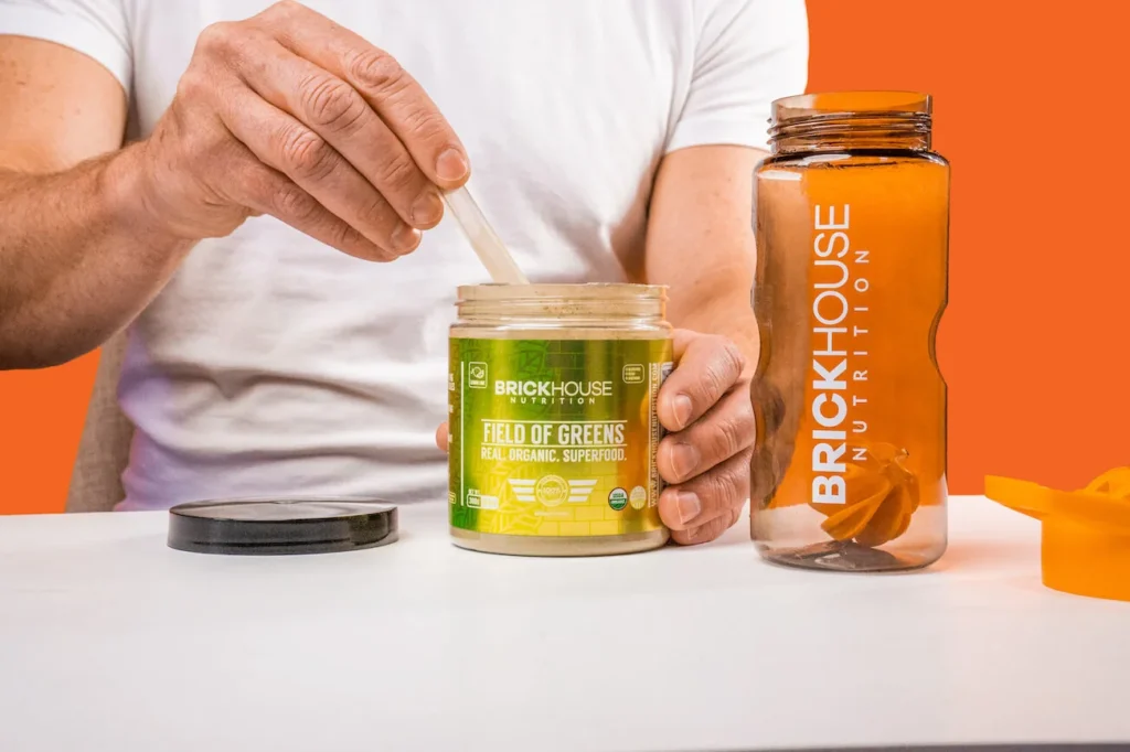 A person's hand stirring a jar of Field of Greens next to a water bottle.