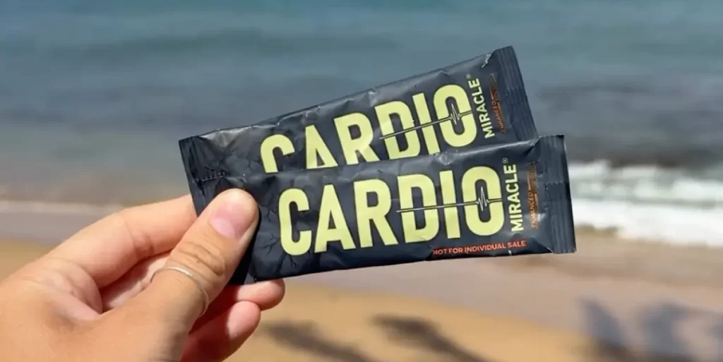 A hand holding two packets of Cardio Miracle supplement on the beach.