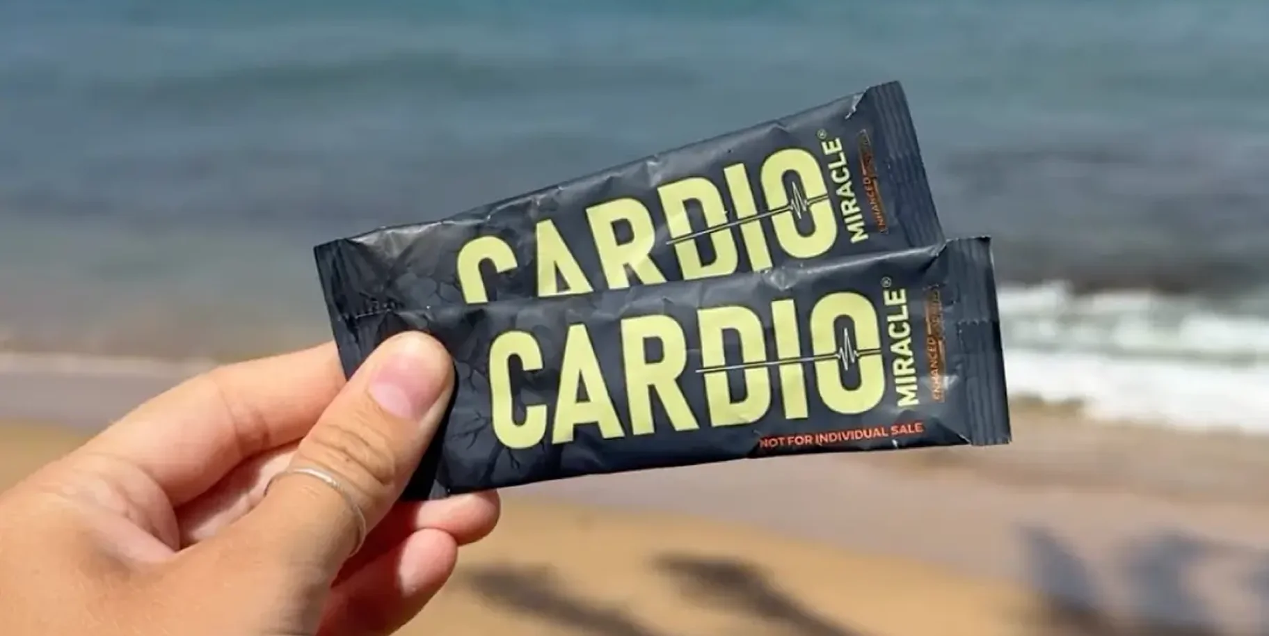 A hand holding two packets of Cardio Miracle supplement on the beach.