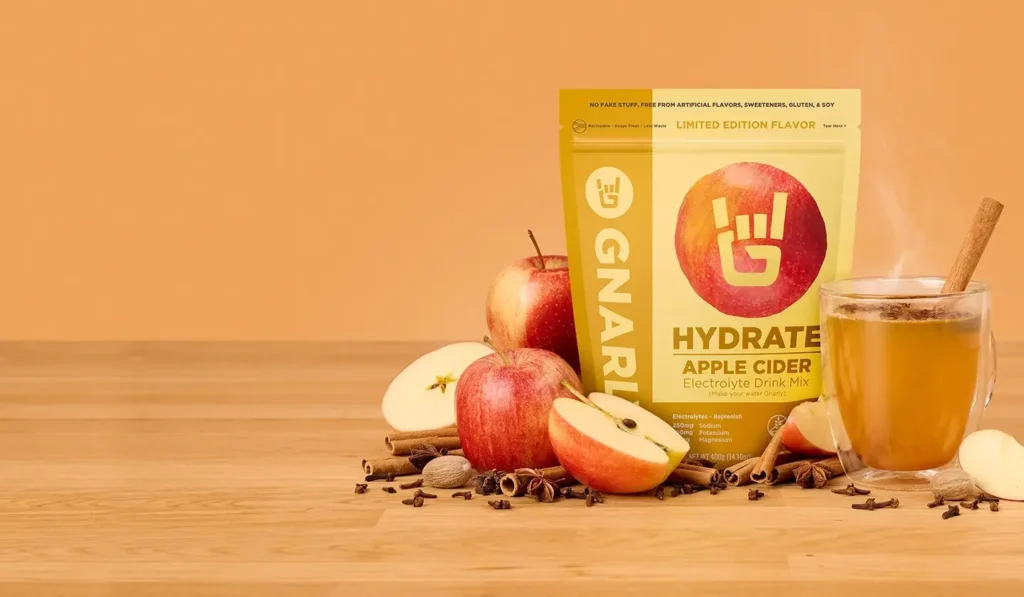 A packaging of Gnarly Hydrate Apple Cider Electrolyte Drink Mix alongside fresh apples and a glass of apple cider.
