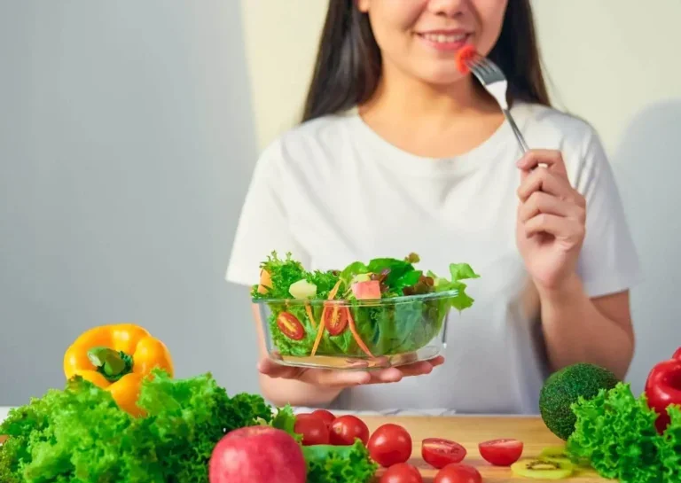 A person holding a bowl of salad while smiling and preparing to eat a piece of tomato.