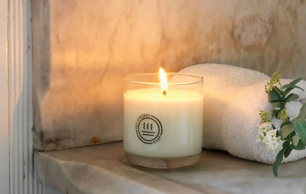 A lit candle sitting on a marble surface next to a rolled towel and foliage.