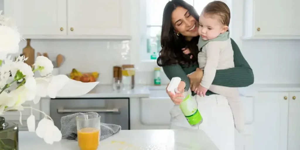 A person holding a child while using a cleaning spray in a kitchen.