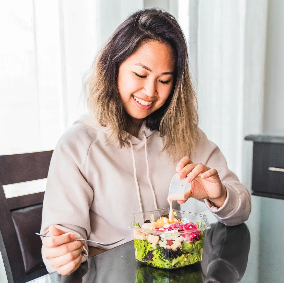 A person pouring dressing onto a salad while sitting at a table.