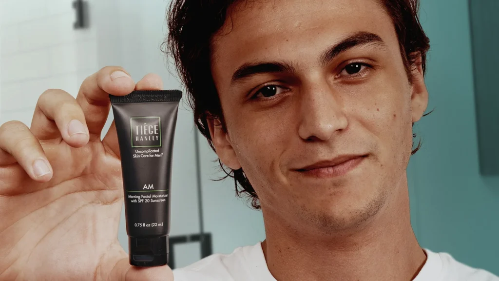 A person holding a Tiege Hanley AM facial moisturizer tube with a smile.