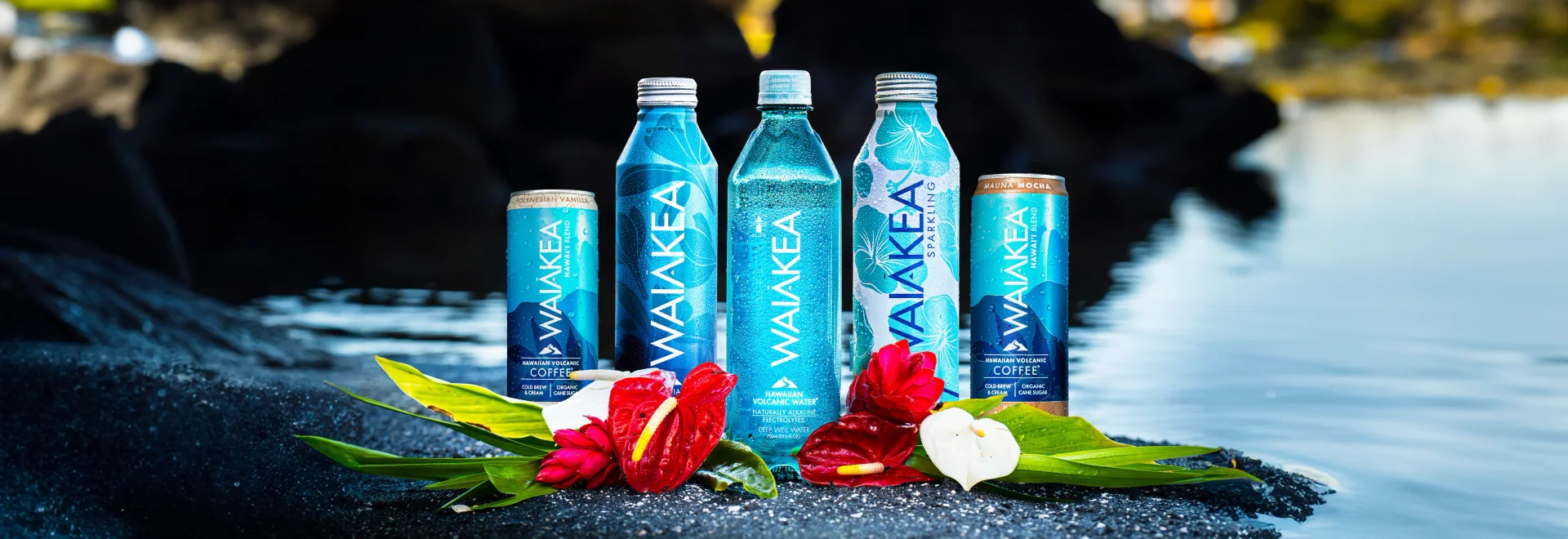 A collection of Waiakea beverages displayed on a rocky surface with tropical flowers.