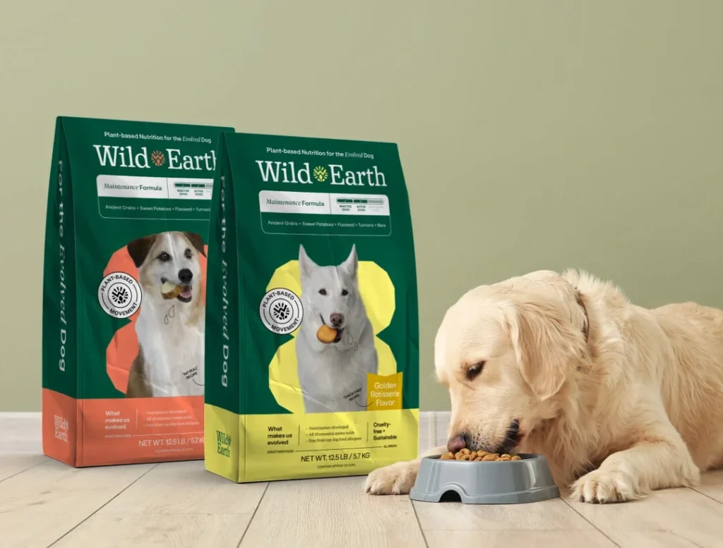 Two bags of Wild Earth Maintenance Formula dog food next to a golden retriever eating from a bowl.