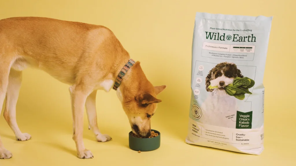 A dog eating from a bowl next to a bag of Wild Earth Performance Formula dog food.