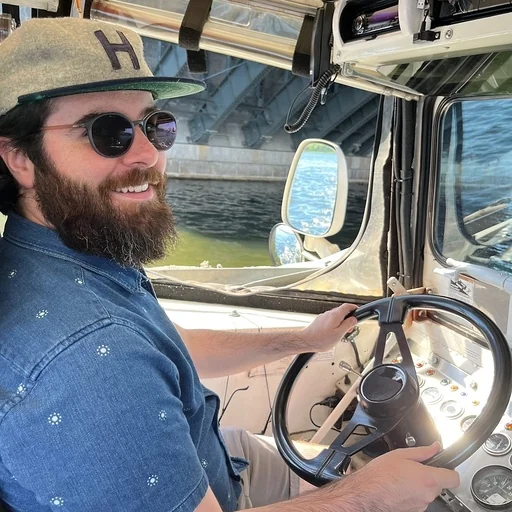 A person with a beard wearing sunglasses and a hat, sitting at the steering wheel of a boat.
