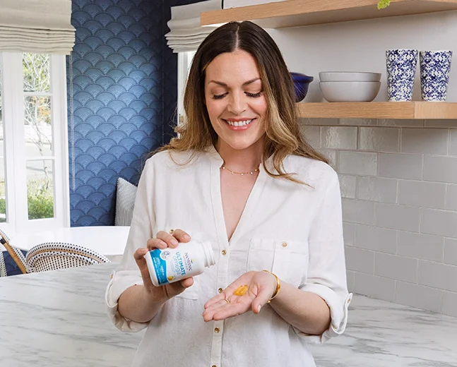 A person holding a bottle of Nécessaire vitamins and a vitamin capsule in their hand in a kitchen setting.