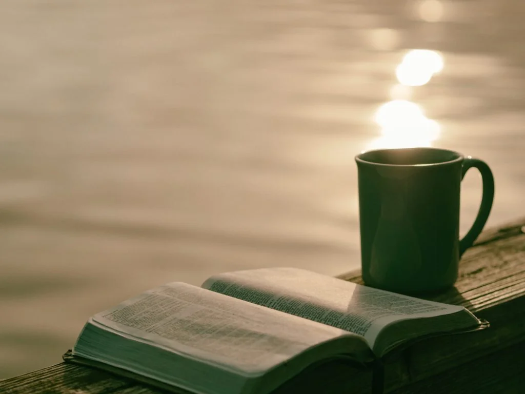 An open book beside a green coffee mug on a wooden surface near water.