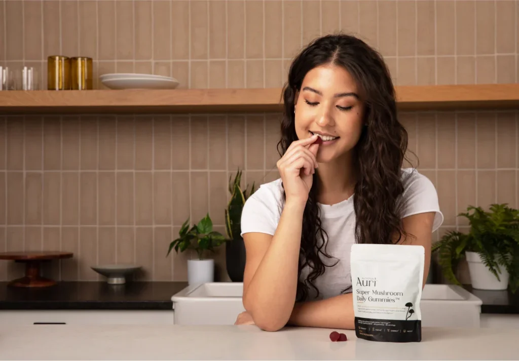 A person smiling while holding a red gummy and sitting near a bag of Auri Super Mushroom Daily Gummies.