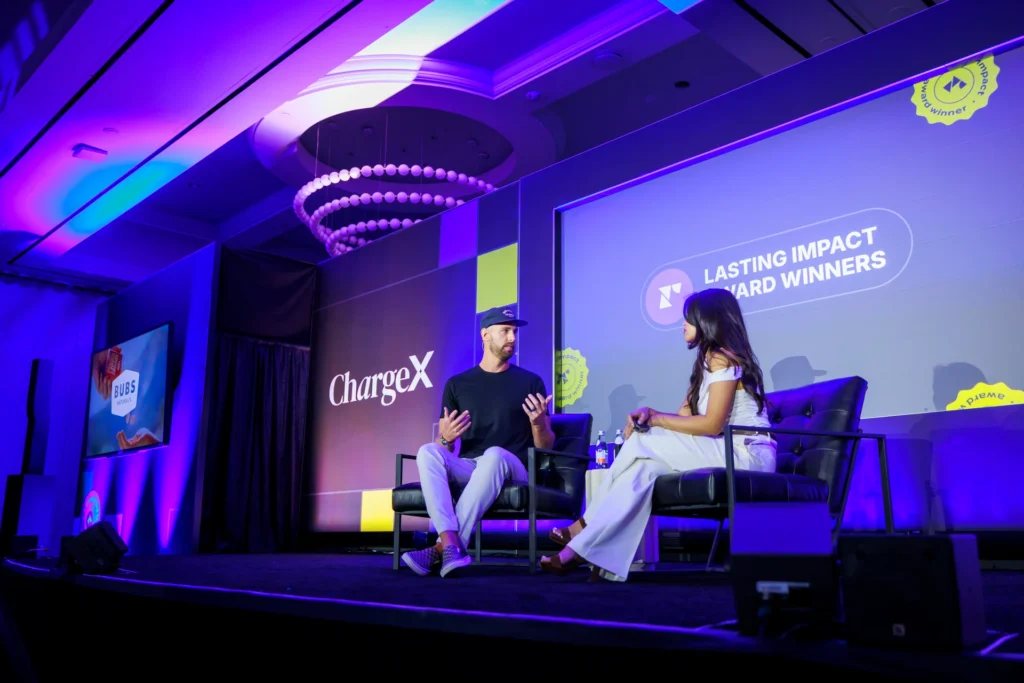 Two speakers discussing at the ChargeX event with Bubs Naturals branding in the background.