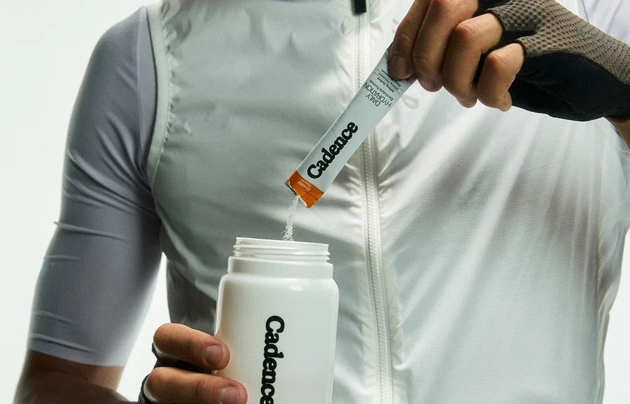 A person pouring powdered drink mix from a tube into a white bottle labeled Cadence.